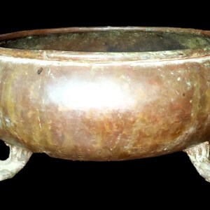 Pode incluir: Uma tigela antiga de bronze, de cor bronze, com corpo arredondado e borda ligeiramente alargada. Assenta em três pés decorativos em forma de garra. O metal tem um aspeto envelhecido com áreas de oxidação verde. O fundo é preto.