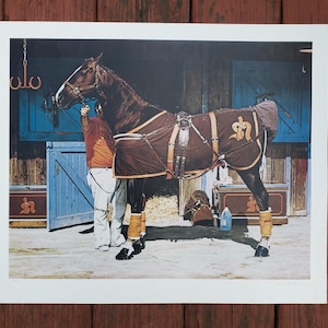 May include: A framed print of a horse in a stable. The horse is dark brown with a blanket and leg wraps. A person in an orange shirt and white pants is adjusting the horse's tack. The stable doors are blue.