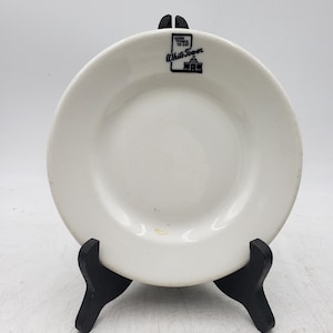 May include: White ceramic plate with the text "Good Things to Eat" and "White Tower" in black. The plate is displayed on a black stand against a white background. The plate has a simple, classic design.