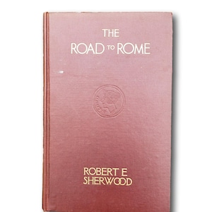 Könnte beinhalten: Ein geschlossenes, rotbraunes Hardcover-Buch mit dem Titel "THE ROAD TO ROME" von Robert E. Sherwood. Der Titel und der Name des Autors sind in Goldbuchstaben gehalten. Auf dem Einband befindet sich ein geprägtes, kreisförmiges Design.