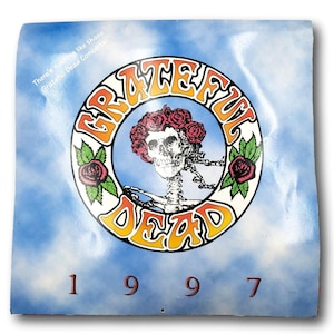 May include: A vintage Grateful Dead concert poster from 1997. The design features a skeleton with a floral crown, surrounded by the band's name in a circular orange and yellow design. The background is a blue sky with clouds.