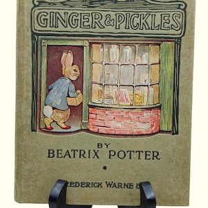 Op de afbeelding: Een hardcover boek getiteld Ginger & Pickles van Beatrix Potter met een beige kaft. De illustratie toont een konijn in een blauw jasje dat een winkel met een bakstenen onderkant en een grote etalage vol glazen potten binnenstapt.
