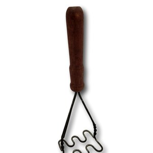 Puede incluir: Un antiguo pasapurés con un mango de madera marrón oscuro y una cabeza de malla ondulada. El alambre metálico es oscuro y el mango muestra signos de desgaste. Esta herramienta de cocina está diseñada para machacar patatas y otros alimentos blandos.