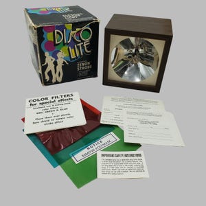 May include: A square Disco Lite Zenon Strobe lamp with a dark wood frame and a circular reflective interior. Includes the original cardboard box, three transparent red, green, and blue color filters, a registration card, and safety instruction sheets.
