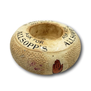 May include: A cream-colored, donut-shaped ceramic ashtray with the text "ASK FOR ALLSOPP'S" in black lettering. The ashtray has a textured surface and a painted orange hand on the side.