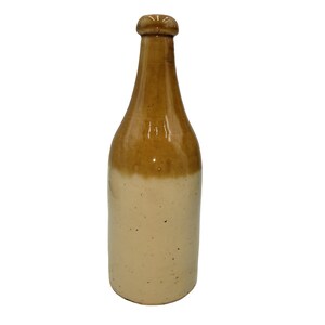 May include: A tall ceramic bottle featuring a two-tone glaze design. The upper portion, including the neck and rounded rim, is finished in a glossy brown glaze, while the lower cylindrical body is finished in a smooth, speckled cream-colored glaze.