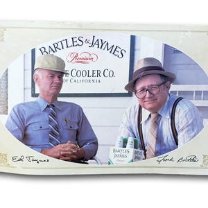 Op de afbeelding: Een ovale vintage advertentie met twee mannen in overhemden en stropdassen, waarvan één met een hoed. Het bord erachter luidt "BARTLES & JAYMES Premium COOLER CO. OF CALIFORNIA." De afbeelding bevat de tekst "Ed Jaymes" en "Frank Bortba" en een sixpack Bartles & Jaymes.