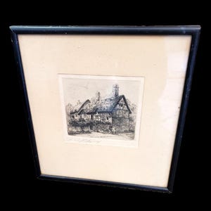 May include: A framed black and white etching depicting a thatched-roof cottage with timber-framed walls and two chimneys. The artwork is set within a cream-colored mat and a thin black frame, featuring a handwritten signature below the image.