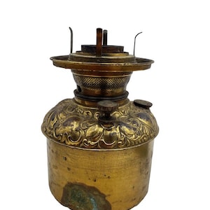 May include: A vintage brass oil lamp base featuring an embossed floral pattern on the shoulder, a cylindrical body with visible patina, and a central burner assembly with three metal prongs and two circular adjustment knobs on the top surface.