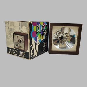 Peut inclure: Un stroboscope carré Disco Lite avec un cadre en bois brun foncé et un intérieur circulaire argenté réfléchissant. La boîte en carton incluse présente le texte Disco Lite, des illustrations de danseurs et des cercles colorés.