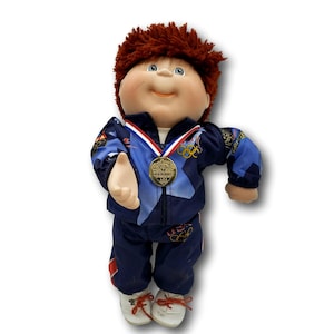 May include: A Cabbage Patch Kids doll in a blue and white Olympic tracksuit and a gold medal. The doll has red hair, blue eyes, and white trainers with red laces. The tracksuit features Olympic rings and USA markings.