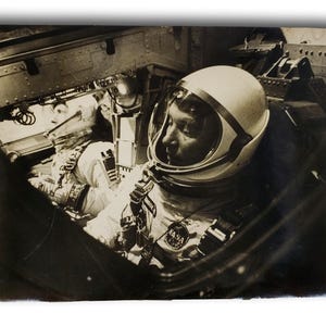 Puede incluir: Fotografía en blanco y negro de un astronauta dentro de una nave espacial. El astronauta lleva un traje espacial y un casco blancos con una visera transparente. El logotipo de la NASA es visible en el traje.