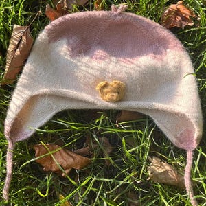 Könnte beinhalten: Eine handgefertigte Ohrenklappenmütze in weichen, gedämpften Rosa- und Cremefarben. Die Mütze hat eine kleine, strukturierte, hellbraune Bärenapplikation auf der Vorderseite. Die Mütze ist aus weichem Strickmaterial gefertigt und hat lange Bänder.