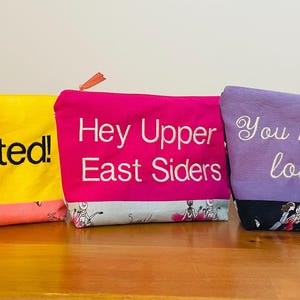 May include: Three zippered pouches with different colored fabric and embroidered text. The pouch on the left is yellow with pink fabric on the bottom and the text "Spotted!" embroidered in black. The middle pouch is pink with a light blue fabric on the bottom and the text "Hey Upper East Siders" embroidered in white. The pouch on the right is purple with black fabric on the bottom and the text "You know you love me" embroidered in white.