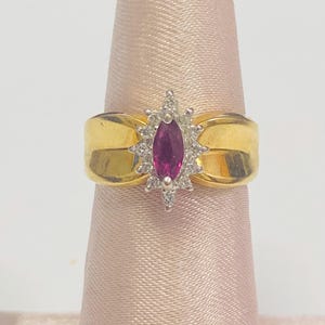 May include: A gold ring featuring a marquise-cut ruby gemstone surrounded by small diamonds. The ring has a wide, polished gold band with a slight curve. The ruby is a deep pink color, and the diamonds sparkle.