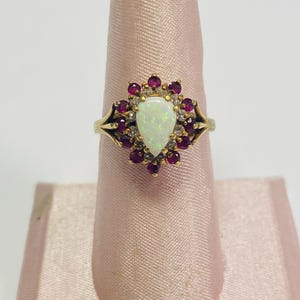 May include: A gold ring featuring a pear-shaped opal center stone, surrounded by small, round, red gemstones and tiny clear stones. The ring is set on a pink display stand.