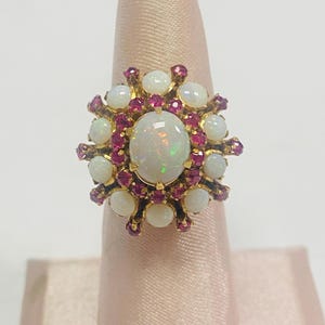 May include: A gold ring featuring a large central oval cabochon opal surrounded by a halo of smaller round rubies and a secondary outer halo of round cabochon opals. The setting is crafted in yellow gold with prong settings for each stone.