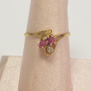 May include: A gold ring featuring three marquise-cut, ruby-colored gemstones and a small diamond accent. The ring has a swirling design and is displayed on a pink fabric background. The ring is a piece of jewelry.
