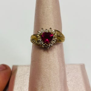 May include: A gold-toned ring featuring a heart-shaped, deep red gemstone at its center, surrounded by smaller, clear stones. The ring's band has a textured design. The ring is displayed on a pink fabric.
