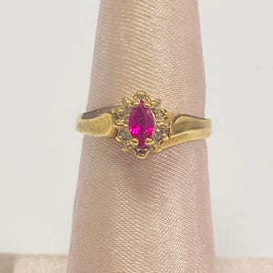 May include: A gold ring featuring a marquise-cut, vibrant pink gemstone, surrounded by smaller, clear stones. The ring has a classic design with a slightly curved band. The ring is displayed on a pink fabric surface.