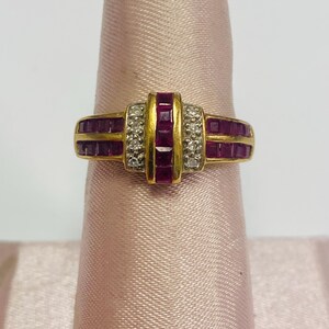 May include: A gold ring featuring a central band of square-cut rubies flanked by rows of smaller rubies and diamond accents. The ring is displayed on a pink surface, showcasing its intricate design and vibrant colors.