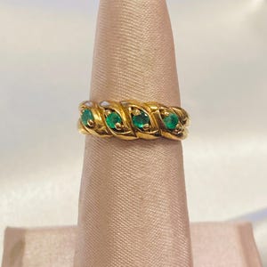 May include: A gold band ring featuring a twisted rope-like design with four small, round, green emerald gemstones set evenly across the front. The metal has a polished finish, and the stones are secured within the textured, braided gold setting.