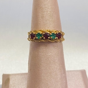 May include: A gold ring featuring a braided band design with five alternating round gemstones. The stones consist of three deep red rubies and two vibrant green emeralds set in a row between the twisted gold metal edges of the ring band.