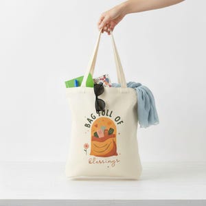 May include: Cream-colored tote bag with the words "BAG FULL OF BLESSINGS" printed on the front. The bag features a graphic of a bag overflowing with items. Sunglasses and a blue scarf are visible inside the bag.