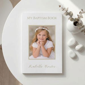 May include: A white baptism book with the text "MY BAPTISM BOOK" and "Isabella Carter" in gold script. The book features a framed photograph of a young girl wearing a floral crown. A white vase with greenery and white stones are on the table.