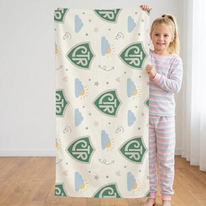 May include: A cream-colored blanket with a repeating pattern of green shield emblems, clouds, suns, stars, and arrows. The blanket is held by a person wearing striped pajamas.