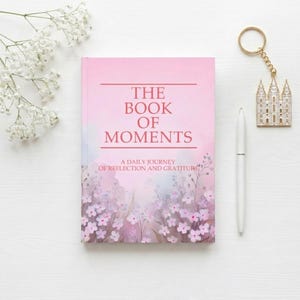 Può includere: Un libro rosa intitolato THE BOOK OF MOMENTS A DAILY JOURNEY OF REFLECTION AND GRATITUDE con un'illustrazione floreale sul fondo, accompagnato da una penna bianca e un portachiavi in metallo dorato con la sagoma di un edificio.