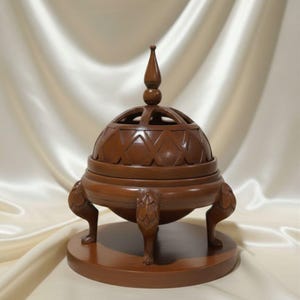May include: A brown wooden incense burner with a domed lid featuring geometric cutouts and a pointed finial. The burner rests on three carved feet and a round base, showcasing intricate craftsmanship.