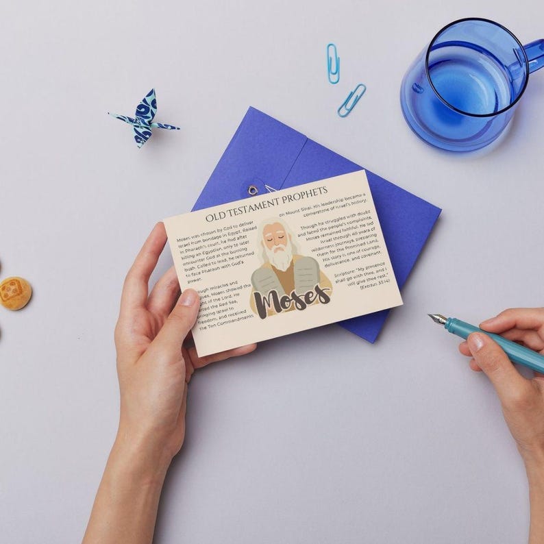 May include: A greeting card with the text "Old Testament Prophets" and an illustration of Moses. The card is held in front of a blue envelope. A blue origami bird, a glass of water, and a pen are also visible.