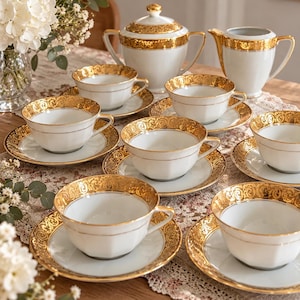 Pode incluir: Um conjunto de chá em porcelana branca com seis chávenas e pires, um açucareiro com tampa e uma leiteira. Cada peça é decorada com uma larga faixa de filigrana dourada nas bordas, complementada por finas linhas douradas na cerâmica branca.