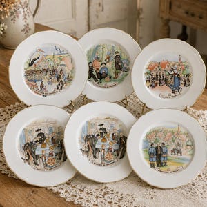 Può includere: Un set di sei piatti in ceramica bianca con bordi smerlati e finiture dorate, ognuno caratterizzato da un'illustrazione colorata di vita di villaggio e didascalie in francese come Le Retour des Cigognes e La Brasserie du Village.