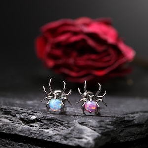 Puede incluir: Dos pendientes con forma de araña con centros de ópalo. Uno tiene un centro azul y verde, el otro un centro rosa y morado. Los pendientes son plateados y están sobre un fondo oscuro con una rosa roja borrosa.