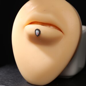 Könnte beinhalten: Nahaufnahme eines Lippenpiercings. Der Piercing-Schmuck hat einen tropfenförmigen Opalstein, der von einer dunklen, perlenbesetzten Fassung umgeben ist. Der Opal hat eine weiße und hellblaue Farbe. Der Piercing-Schmuck befindet sich auf einer Schaufensterpuppe.