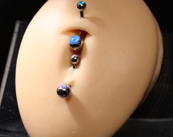 May include: Close-up of a belly button with multiple belly button rings. The rings have a black post and a round, iridescent gem. The gems have a blue, purple, and pink color scheme. The skin tone is light.