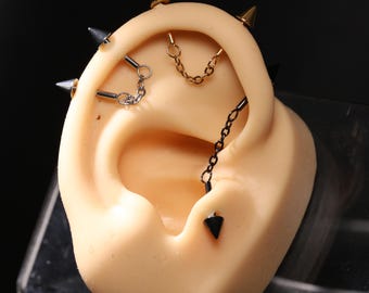 May include: An ear model showcasing various earrings. The ear features chain and spike earrings in silver, gold, and black. The jewelry creates a bold, contemporary look. The image highlights the different styles and colours of the earrings.