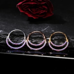 Op de afbeelding: Drie oorringen met een dubbellaags opaalontwerp. De oorbellen zijn in zilver, roségoud en goudtinten. Een donkere achtergrond en een rode roos zijn zichtbaar.