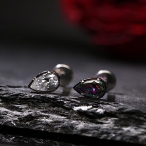 May include: Two silver stud earrings with teardrop-shaped gemstones. One earring features a clear, faceted stone, while the other has a shimmering, iridescent gem with a range of colors. The earrings are set against a dark, textured background.