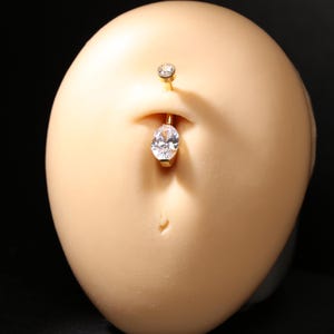 May include: A gold-colored belly button ring with a clear, oval-shaped gem hanging from the bottom and a smaller, round gem on top. The jewelry is set against a light skin-toned background.