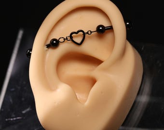 May include: A black industrial barbell piercing jewelry featuring a central open heart charm connected by small links, flanked by two smooth black spherical beads on each side, positioned horizontally across the upper cartilage of an ear model.
