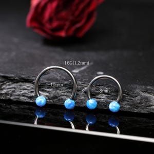 May include: Two horseshoe-shaped, silver-colored body jewelry pieces with blue opal-like beads. One is 10mm in diameter, the other is 8mm. The gauge is 16G (1.2mm). The jewelry is displayed on a dark surface.