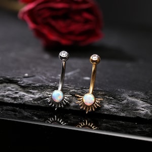 Pode incluir: Dois piercings para o umbigo. Um é prateado, o outro dourado. Cada um apresenta uma pequena pedra opalescente redonda na parte inferior, rodeada por raios em forma de sol. Uma gema transparente está no topo de cada anel. O fundo é escuro.
