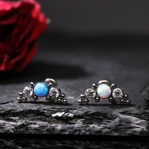 Könnte beinhalten: Zwei silberfarbene, gebogene Ohrringe. Jeder Ohrring hat einen zentralen, runden, blauen Opalstein, flankiert von klaren, runden Kristallen und kleinen, silberfarbenen Perlen. Die Ohrringe werden auf einer dunklen, strukturierten Oberfläche präsentiert.