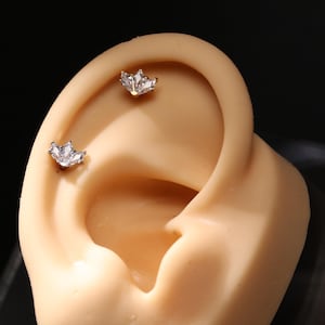 May include: Two silver-toned, crown-shaped earrings with clear, sparkling stones are displayed on a tan ear. The earrings are small and delicate, with a cluster of stones forming the crown design. The background is dark.