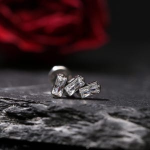 May include: A silver-toned earring with three rectangular, clear crystals. The earring is set against a dark, textured surface with a blurred red rose in the background. The crystals are arranged in a cluster.