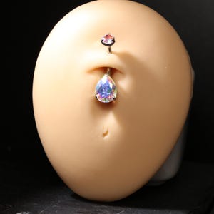 May include: A belly button ring with a teardrop-shaped, iridescent gem. The top gem is small and pink. The jewelry is silver-toned and is displayed on a light-colored, oval-shaped form.
