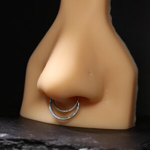 May include: A silver septum ring with a double hoop design, featuring a white opal-like inlay. The jewellery is displayed on a mannequin nose against a dark background, showcasing the piercing.